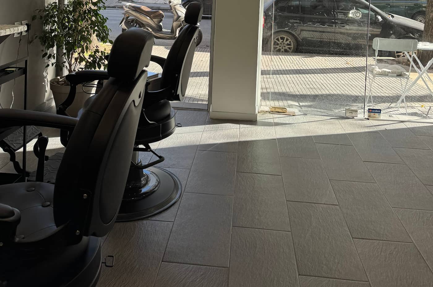 Οι δύο μαύρες καρέκλες του GOSSHEEP BARBERSHOP και η τζαμαρία προς το δρόμο, με το φως της μέρας πάνω στα πλακάκια